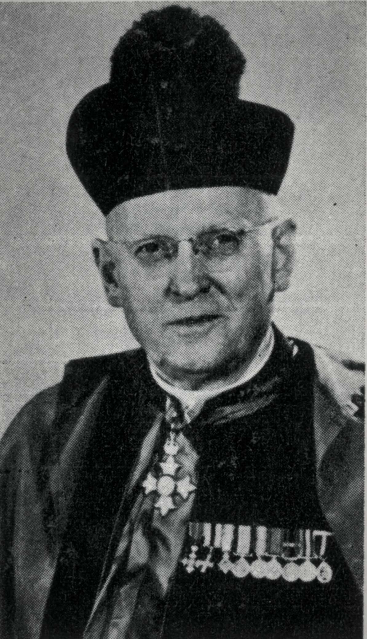 Portrait en noir et blanc – Un homme âgé est photographié à partir de la poitrine dans son habit religieux, y compris un chapeau et une cape. Il porte des médailles militaires miniatures et un insigne en sautoir de l’Ordre de l'Empire britannique.